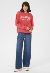 Sweatshirt rouge avec "LOS ANGELES Dreamers Club" imprimé en blanc, associé à un jean denim bleu ample et des baskets blanches.