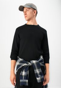 Jeune homme portant une casquette grise, une chemise à manches longues noire et une chemise à carreaux nouée autour de la taille, regardant vers la gauche devant un fond blanc.