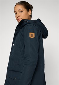 Marineblauwe parka jas met een gevoerde capuchon, voorzien van een bruine logo patch, drukknoopsluiting en zijzakken voor functionaliteit. Soepele textuur.