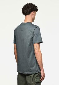 Joven con cabello rizado, vistiendo una camiseta gris de manga corta y pantalones cargo verdes, de pie mirando hacia atrás en un fondo plano.