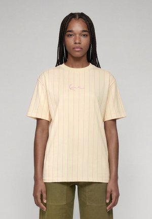 Jeune femme aux cheveux tressés portant une chemise à manches courtes jaune pâle avec de fines rayures verticales rouges et un pantalon vert olive, debout devant un fond gris.
