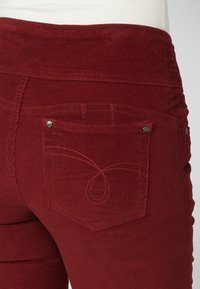 Gros plan sur la taille et la poche arrière d'un pantalon en velours côtelé rouge avec coutures décoratives et rivets métalliques.