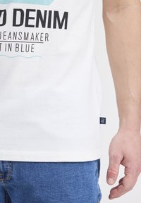 Tricou alb cu grafică imprimată "DENIM", accente albastre și etichetă logo pe lateral; purtat cu blugi din denim albastru. Textura netedă a țesăturii din bumbac.