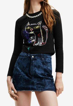 Femme portant un haut noir à manches longues avec un design de chat coloré en contour et une mini-jupe en denim bleu foncé à motifs.