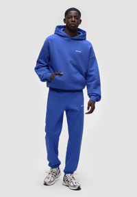 Felpa blu con tasca a marsupio e pantaloni joggers blu abbinati. Presenta un logo bianco sul petto e sulla gamba. Il modello indossa scarpe da ginnastica argentate.