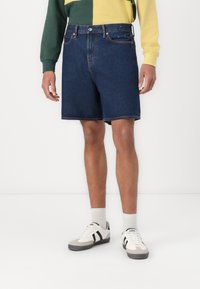 Denim shorts in dark blue with a straight cut, five pockets, and visible stitching. Paired with white and gray sneakers and light socks.