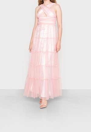 Vrouw draagt een mouwloze, tot op de grond reikende lichtroze jurk met een kruisvormig lijfje en laagjes transparante stof, gecombineerd met nude open-toe hakken.