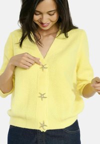 Vrouw in een gele, gestructureerde cardigan met zeester-vormige knopen die het sluit, gecombineerd met donkerblauwe jeans.