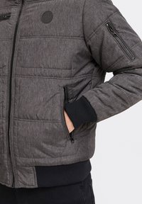 Padded jacket in dark gray woven fabric, featuring a zip front, ribbed cuffs, and multiple zip pockets for storage. Textured detailing visible.