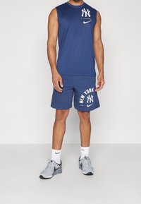 Canotta blu navy con logo NY sul petto, abbinata a pantaloni corti coordinati con stampa "NEW YORK" e logo. Sneakers grigie.