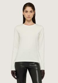 Blusa blanca de manga larga hecha de tela suave con un escote redondo, combinada con pantalones de cuero negro brillantes y guantes oscuros, contra un fondo neutro.