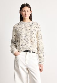 Grober cremiger Pullover mit strukturierter Strickoptik und blauen Sprenkeln. Wird über weißen Hosen getragen, mit einem schwarzen Gürtel und einem Rundhalsausschnitt.