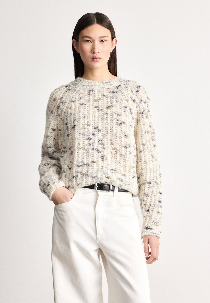 Grober cremiger Pullover mit strukturierter Strickoptik und blauen Sprenkeln. Wird über weißen Hosen getragen, mit einem schwarzen Gürtel und einem Rundhalsausschnitt.