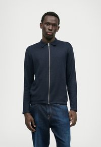 Suéter de polo de malha azul-marinho com fecho zip, colarinho, mangas compridas e bainha canelada, combinado com jeans de ganga azuis. Textura suave, design minimalista.