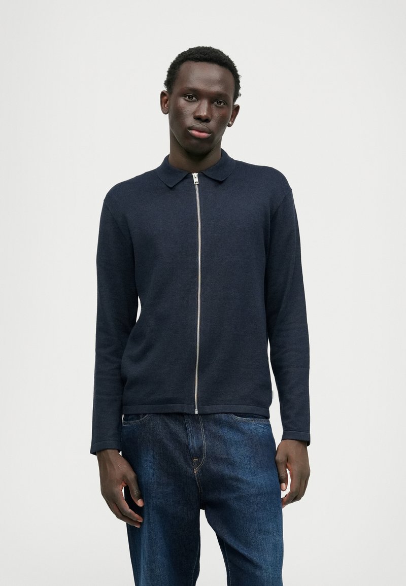 Suéter de polo de malha azul-marinho com fecho zip, colarinho, mangas compridas e bainha canelada, combinado com jeans de ganga azuis. Textura suave, design minimalista.