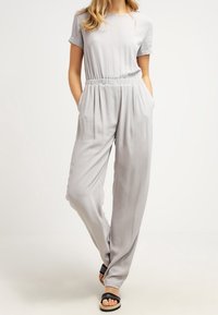 Jasnoszary kombinezon z krótkimi rękawami, elastycznym pasem, luźnym krojem i bocznymi kieszeniami. Gładka tekstura z płynnie opadającym fasonem i minimalistycznym designem.