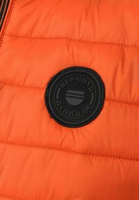 Segment van een oranje gewatteerde jas met een zwart rubberen rond logo met reliëftekst en een verhoogd cirkelvormig ontwerp. Gentextureerd, geborduurd oppervlak.