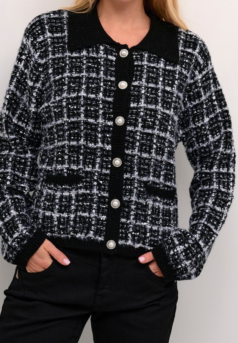 Femme portant un cardigan en maille texturée noir et blanc avec des boutons en perle et deux poches à l'avant, assorti à un pantalon noir.