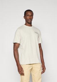 Ανοιχτό καφέ βαμβακερό T-shirt με κοντά μανίκια, στρογγυλή λαιμόκοψη και ένα μικρό μαύρο λογότυπο στο αριστερό στήθος. Το μοντέλο φοράει ανοιχτό καφέ παντελόνι.