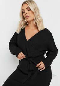 Musta kietaisumekko pehmeästä neuloksesta, jossa on V-pääntie, pitkät hihat ja solmittava vyötäröyksityiskohta säädettävää istuvuutta varten. Sujuva pinta.