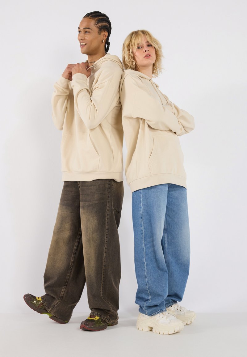 Beige hoodies with front pockets, worn by two individuals. One in dark denim, the other in light blue jeans. Sneakers with thick soles visible.