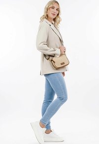 Blazer beige sobre una blusa blanca, jeans de mezclilla azul claros y zapatillas blancas. Sostiene un bolso cruzado beige con textura tejida y acentos dorados.