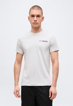 Uomo che indossa una maglietta Lacoste grigio chiaro e pantaloncini neri, in piedi davanti a uno sfondo chiaro e uniforme, con espressione neutra e volto frontale.