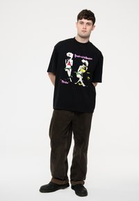 Black oversized t-shirt featuring a colorful graphic print of two figures. Paired with brown loose-fitting pants and black shoes.