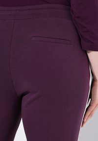 Close-up van een persoon in paarse broek met een achterzak en een witte zijstreep, hand zachtjes rustend op de heup.