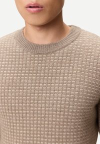 Beige stickad tröja med ett strukturerat, ribbat mönster och en klassisk rund halsringning, tillverkad av ett mjukt, mediumtungt material.