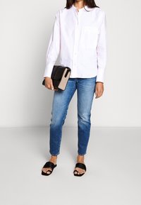 Vit skjorta med knapp-up och ficka, tillsammans med ljusblå jeans och svarta slidesandaler. En svart och beige handväska hålls i handen.