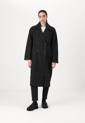 Cappotto doppiopetto in lana nera con ampi rever a punta, tasche laterali e maniche lunghe, indossato sopra jeans scuri e scarpe nere.