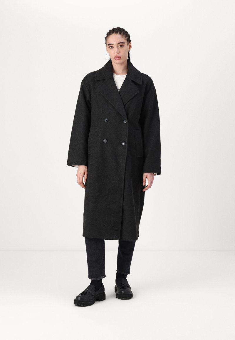 Cappotto doppiopetto in lana nera con ampi rever a punta, tasche laterali e maniche lunghe, indossato sopra jeans scuri e scarpe nere.