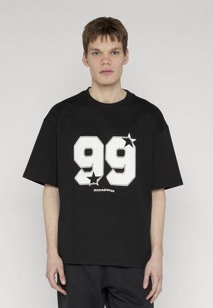 Giovane uomo che indossa una t-shirt nera oversize con il numero bianco "99" e grafiche a stella, in piedi davanti a uno sfondo grigio uniforme.