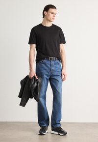 T-shirt en coton noir, jeans en denim bleus, ceinture noire, tenant une veste en cuir noire, portant des baskets noir et blanc, fond neutre.