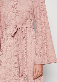 Różowa koronkowa sukienka z dzwonowymi rękawami i pasującym paskiem w talii. Cechuje się kwiatowym wzorem koronkowym na całej tkaninie.