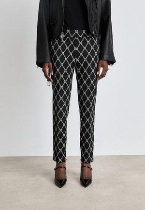 Personne portant un pantalon slim à motif losange noir et blanc, une veste en cuir noire et des talons hauts noirs pointus avec des brides à la cheville.
