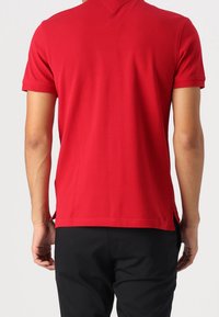 Homme portant un polo rouge uni à manches courtes et un pantalon noir, vu de dos sur un fond clair uni.