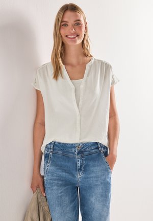 Femme souriante portant un chemisier blanc à manches courtes boutonné et un jean bleu, tenant une veste beige, debout contre un mur blanc.