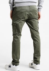 Olivgrüne Cargo-Hosen mit schmalem Schnitt, aus einem Baumwoll-Mischgewebe gefertigt, mit mehreren Taschen und einer glatten Textur. Getragen mit weißen Sneakers.