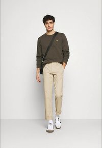 Brun genser med en gul logo, beige bukser og hvite joggesko med brune accenter. Modellen bærer en svart crossover-veske.