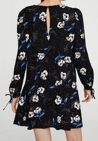 Robe noire à manches longues, motif floral bleu et blanc, détail dos avec échancrure en forme de clé, poignets noués, silhouette évasée et texture subtile.