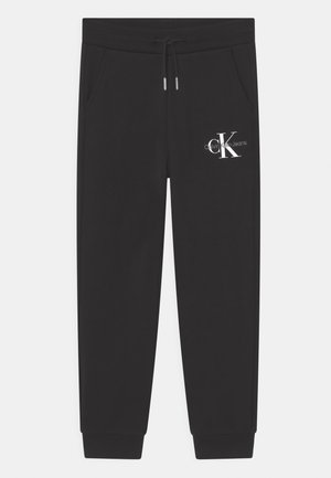 Pantalons de survêtement noirs en tissu doux, avec une taille élastique à cordon, des poches latérales et un logo blanc sur la cuisse gauche. Poignets côtelés aux chevilles.