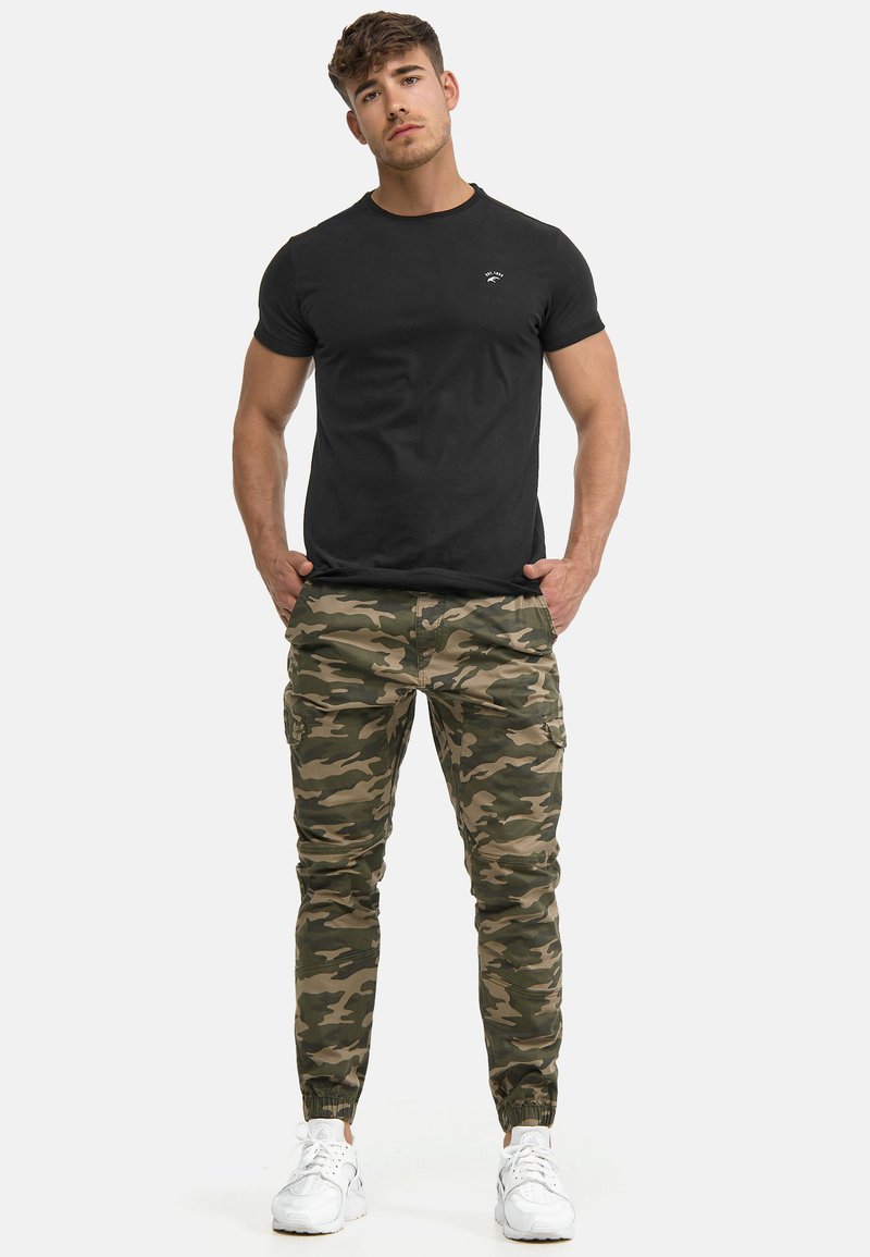 Zwart katoenen T-shirt, getailleerd ontwerp, minimaal logo. Camouflage cargobroek, taps toelopende pasvorm, meerdere zakken, elastische enkelboorden, gedragen met witte sneakers.
