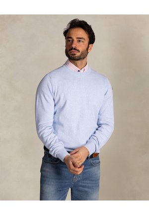 Mann mit dunklem Haar und Bart, der einen hellblauen Pullover über einem rosa Hemd mit Kragen und blaue Jeans trägt, steht mit verschränkten Händen.