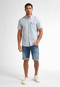 Kortärmlig, lichtblauw gestreept overhemd met een borstzak, gecombineerd met opgerolde denim shorts en gebroken witte sneakers.