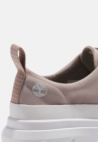 Gros plan sur le talon d'une basket en tissu beige avec une semelle blanche texturée et une languette de traction, arborant un petit logo d'arbre blanc sur le côté.