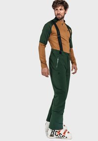 Mann in grünen Skihosen mit schwarzen Hosenträgern, einem braun-grünen Langarmoberteil und weißen Skistiefeln mit orangefarbenen Akzenten.