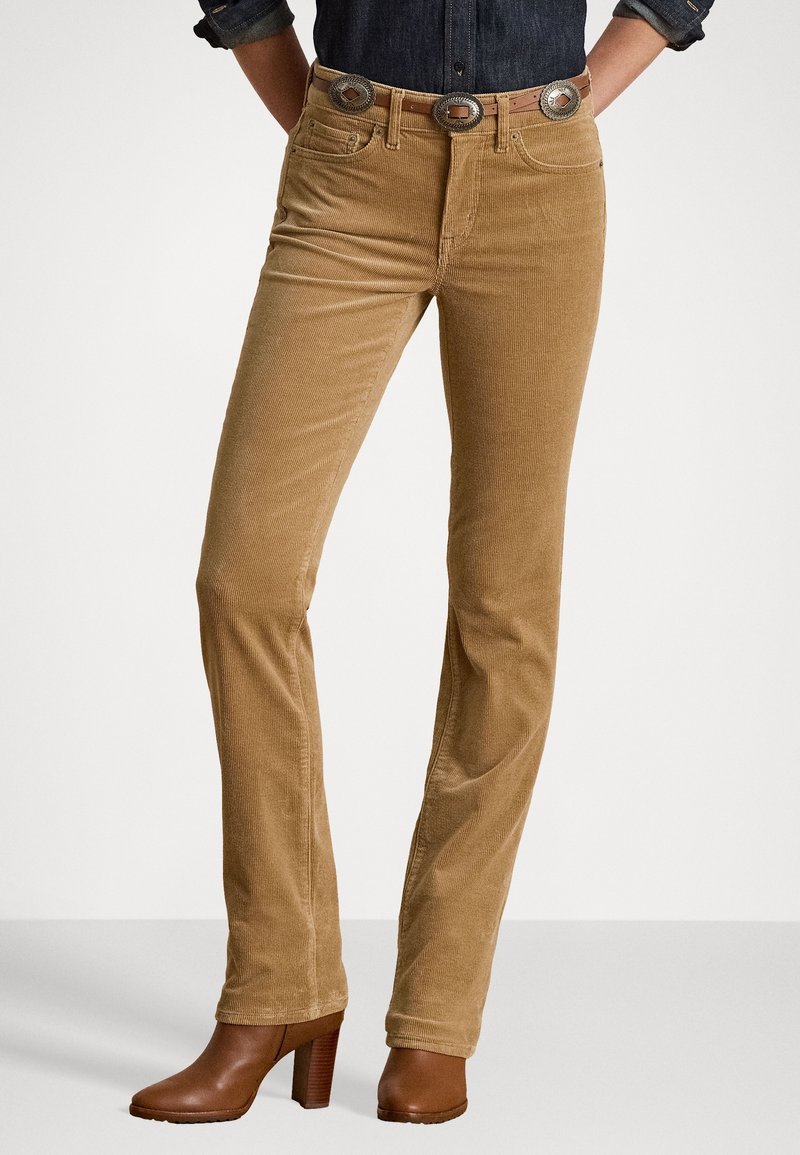 Pantalon en velours côtelé de couleur brun clair avec une coupe slim, présentant une taille haute et des éléments décoratifs en métal argenté sur la ceinture.