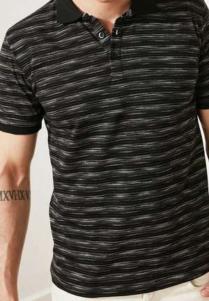 Hombre que lleva una camisa polo de rayas horizontales negras y grises con cuello negro y pantalones beige, con un tatuaje de número romano en el antebrazo izquierdo.